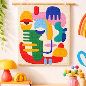 Puede incluir: Tapiz de pared abstracto con un diseño de rostro colorido con formas amarillas, azules, verdes, rojas y rosas. La obra de arte está montada en una barra de madera. Una lámpara amarilla y otros objetos decorativos están sobre una superficie de madera.