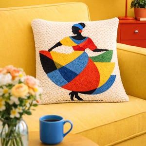 Puede incluir: Un cojín decorativo con un diseño bordado colorido de una figura bailando. La silueta lleva un vestido amarillo, rojo, azul y negro. El cojín está sobre un sofá amarillo, con una lámpara roja y una taza azul cerca.