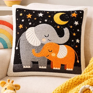 May include: A square decorative pillow featuring a gray elephant and an orange elephant, set against a black background with stars and a crescent moon. The pillow has a white border and a textured appearance, suggesting a handmade or embroidered design.