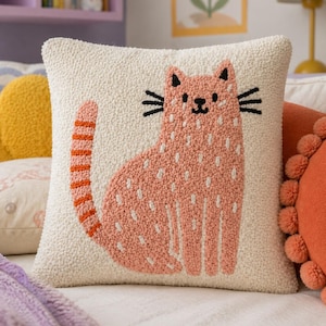 May include: Square off-white cushion with a textured surface. The cushion features a pink cat design with black whiskers and orange stripes on the tail. The cat is embroidered with white dots and lines.
