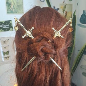 May include: Gold-coloured hair accessories, including sword-shaped hairpins and decorative charms, are styled in red hair. The text "Mystical Bunny" is visible in the background. The hair is styled in a bun.