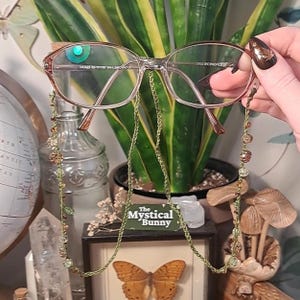 May include: Clear-framed spectacles with a green beaded chain. The chain has small, decorative beads in shades of green, brown, and gold. The spectacles are held in front of a potted plant and a framed butterfly display. The text "The Mystical Bunny" is visible.