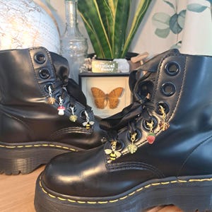 May include: Black combat boots with decorative charms. The boots have black ribbon laces and gold-colored charms, including a bee, a strawberry, and a mushroom. The boots are on a platform sole.