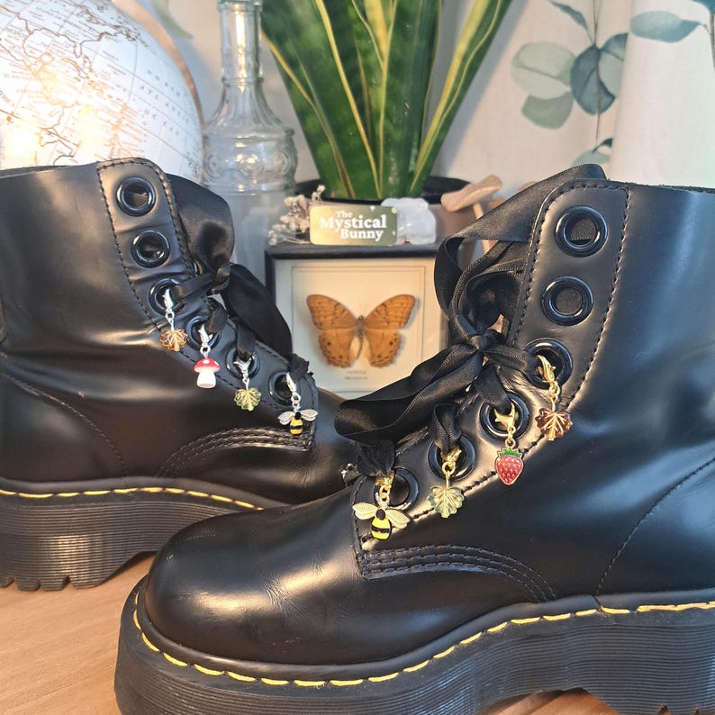 May include: Black combat boots with decorative charms. The boots have black ribbon laces and gold-colored charms, including a bee, a strawberry, and a mushroom. The boots are on a platform sole.