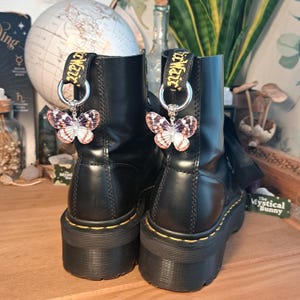 May include: Black leather boots with butterfly charms. The charms are attached to the boot loops with silver rings. The butterfly wings are a mix of white, pink, and purple. The boots have yellow stitching and thick soles.
