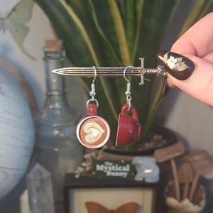 May include: Red coffee cup earrings, one with a heart-shaped latte art design, suspended from a silver sword-shaped earring holder. The background includes a globe, a bottle, and a plant.