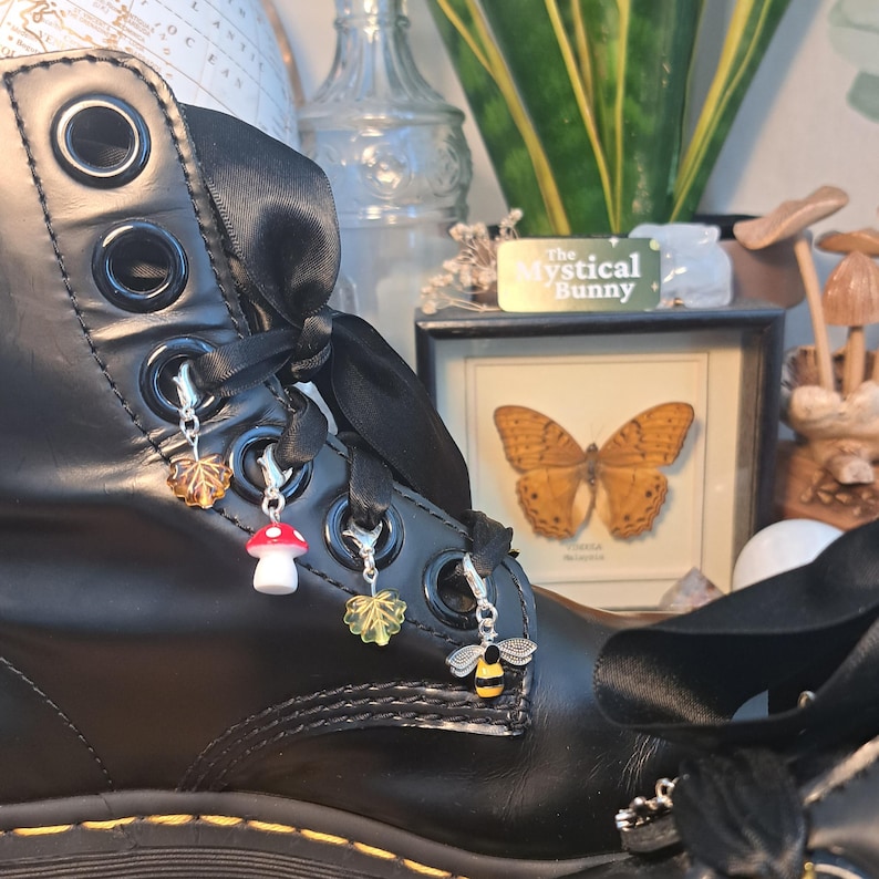 May include: Black leather boot with decorative charms. Charms include a leaf, mushroom, leaf, and bee. The boot has black ribbon laces and silver eyelets. A framed butterfly and a sign that says "The Mystical Bunny" are in the background.
