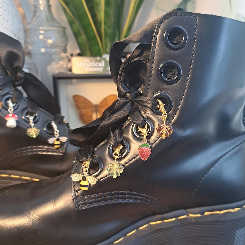 May include: Black leather boots with black ribbon laces and decorative charms. Charms include a red strawberry, a yellow and black bee, a green leaf, and a mushroom. The boots have large eyelets and yellow stitching.