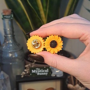 May include: Two sunflower-shaped enamel pins. One pin is a solid yellow with a gold pin back. The other pin has yellow petals and a black center. The pins are held between fingers.