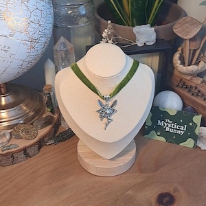 K&ouml;nnte beinhalten: Eine silberne Feen-Anh&auml;nger-Kette mit einem gr&uuml;nen Samt-Choker. Die Kette wird auf einer wei&szlig;en Mannequin-B&uuml;ste auf einem Holzsockel pr&auml;sentiert. Der Text "The Mystical Bunny" ist auf einer gr&uuml;n-wei&szlig;en Visitenkarte sichtbar.