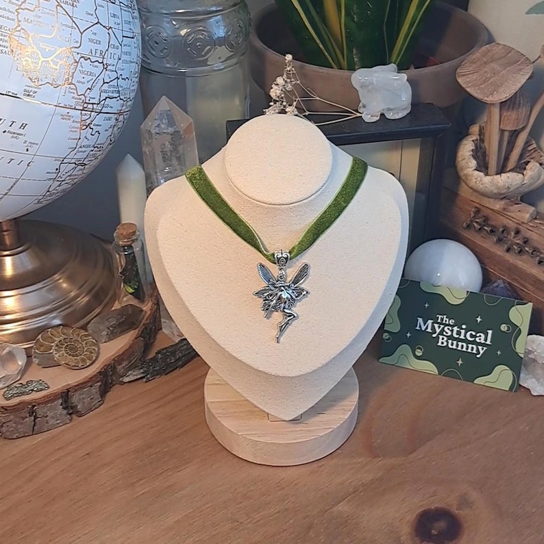 K&ouml;nnte beinhalten: Eine silberne Feen-Anh&auml;nger-Kette mit einem gr&uuml;nen Samt-Choker. Die Kette wird auf einer wei&szlig;en Mannequin-B&uuml;ste auf einem Holzsockel pr&auml;sentiert. Der Text "The Mystical Bunny" ist auf einer gr&uuml;n-wei&szlig;en Visitenkarte sichtbar.