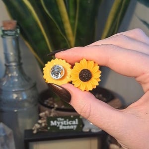 May include: Two sunflower-shaped enamel pins. One pin is a full sunflower with yellow petals and a dark brown center. The other pin is a yellow circle with a silver pin back. The pins are held in front of a green plant.