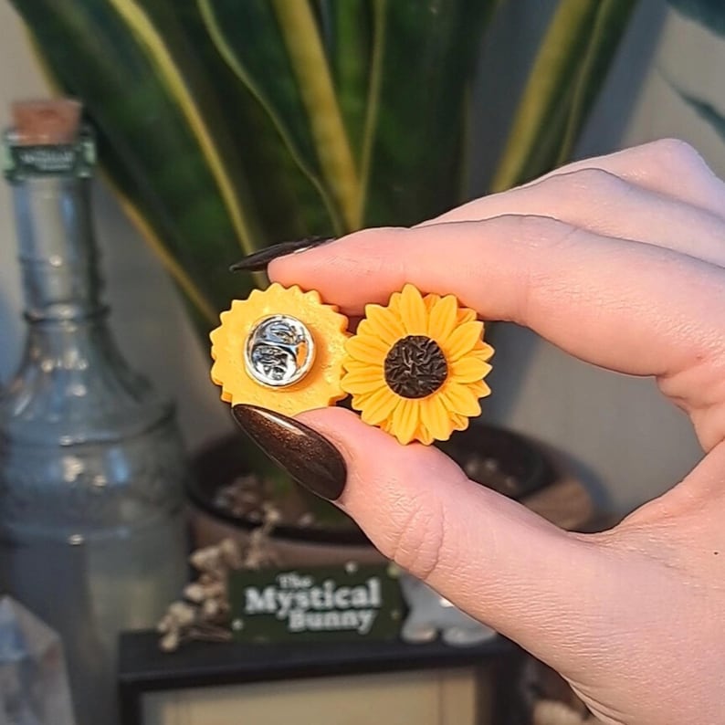 May include: Two sunflower-shaped enamel pins. One pin is a full sunflower with yellow petals and a dark brown center. The other pin is a yellow circle with a silver pin back. The pins are held in front of a green plant.