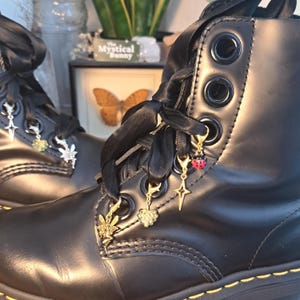 May include: Black leather boots with black ribbon laces and decorative charms. Charms include a fairy, a leaf, a star, and a ladybug. The boots have large eyelets and a stitched sole.