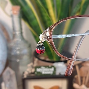 May include: Eyeglasses with a small ladybug charm hanging from the frame. The charm is red with black spots. The glasses have a light brown frame and clear lenses. The background is blurred, with a plant and other objects visible.