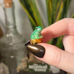 May include: A small, three-dimensional green frog pin featuring a smooth, glossy finish and black eyes. The frog is attached to a circular gold-toned metal backing with a pin post, designed to be secured with a matching gold-toned metal clutch.