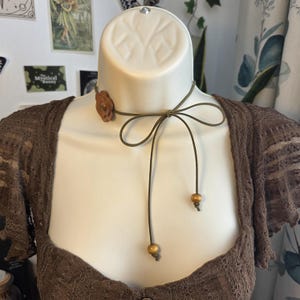 May include: A brown choker necklace with a flower accent and a tie closure. The necklace has a thin, olive green cord with two small wooden beads. The flower is a darker brown and is on the side of the neck.