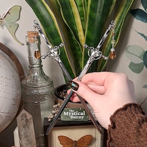 May include: Two silver hair sticks in a sword design, crossed in front of a plant. Each sword has a heart-shaped hilt and a decorative charm with amber-coloured beads. The background includes a globe, a glass bottle, and a framed butterfly.