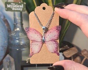 May include: A silver-toned necklace featuring a butterfly pendant with pink and purple wings. The butterfly is attached to a silver chain and displayed on a brown tag. The tag has a hole at the top. The text "The Mystical Bunny" is visible in the background.