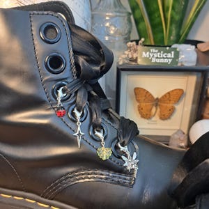 May include: Close-up of a black leather boot with black ribbon laces and decorative charms. Charms include a ladybug, a star, a leaf, and a fairy. The boot has silver eyelets and a black sole. The boot is part of a gothic or alternative fashion theme.