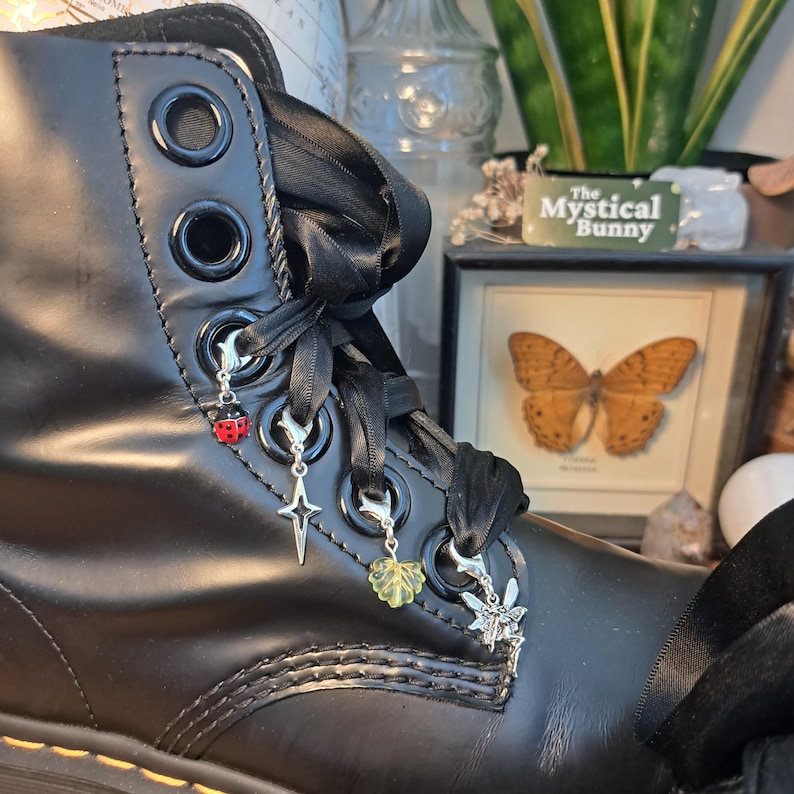 May include: Close-up of a black leather boot with black ribbon laces and decorative charms. Charms include a ladybug, a star, a leaf, and a fairy. The boot has silver eyelets and a black sole. The boot is part of a gothic or alternative fashion theme.