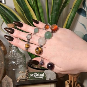 May include: A hand displaying several gemstone rings with silver bands. The rings feature various stones, including tiger's eye, green jade, and amethyst. The nails are painted a dark brown color. The rings are the focal point of the image.