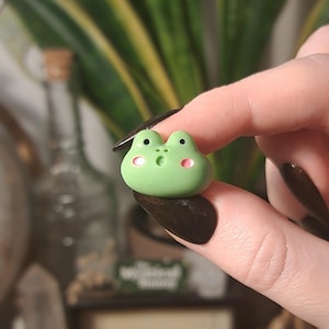 May include: A small, green frog figurine with black eyes and pink cheeks. The frog has a surprised expression. The figurine is held between two fingers. A cute, whimsical design.