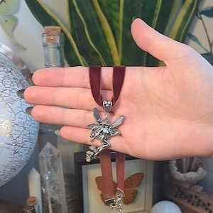 K&ouml;nnte beinhalten: Eine silberne Feen-Anh&auml;nger mit Fl&uuml;geln und einem flie&szlig;enden Kleid h&auml;ngt an einer dunkelroten Samt-Choker-Kette.
