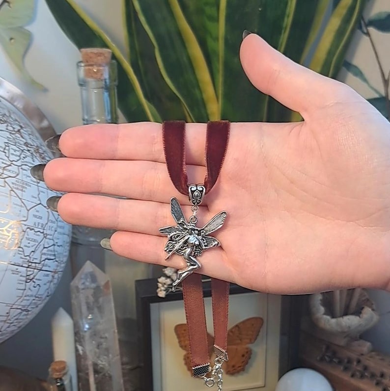 K&ouml;nnte beinhalten: Eine silberne Feen-Anh&auml;nger mit Fl&uuml;geln und einem flie&szlig;enden Kleid h&auml;ngt an einer dunkelroten Samt-Choker-Kette.