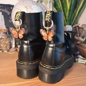 May include: Black platform boots with butterfly charms. The charms are orange and brown with gold-colored chains. The boots have yellow stitching and black pull tabs with the word "Air" in gold lettering.
