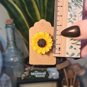 May include: A yellow sunflower brooch with a brown center is attached to a brown tag. A ruler shows the brooch is approximately 1 inch in diameter. The background includes a green plant and a glass bottle.