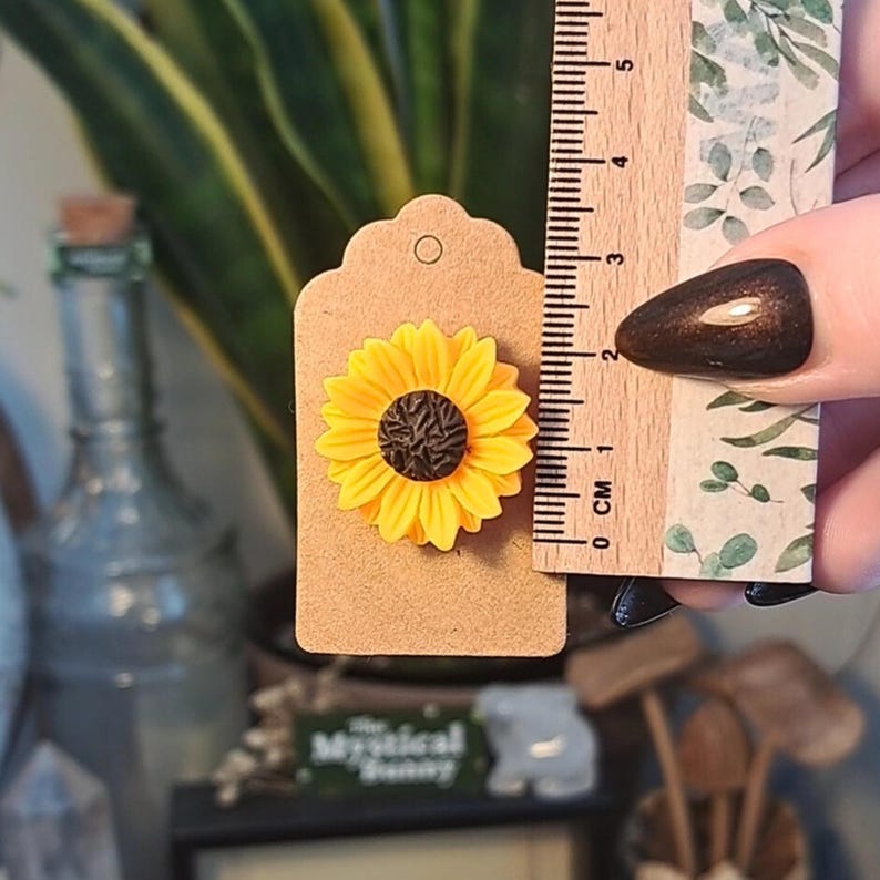 May include: A yellow sunflower brooch with a brown center is attached to a brown tag. A ruler shows the brooch is approximately 1 inch in diameter. The background includes a green plant and a glass bottle.