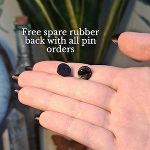 May include: Close-up of two pin backs, one black rubber and one metal. The text "Free spare rubber back with all pin orders" is visible in white. The items are displayed on a hand.