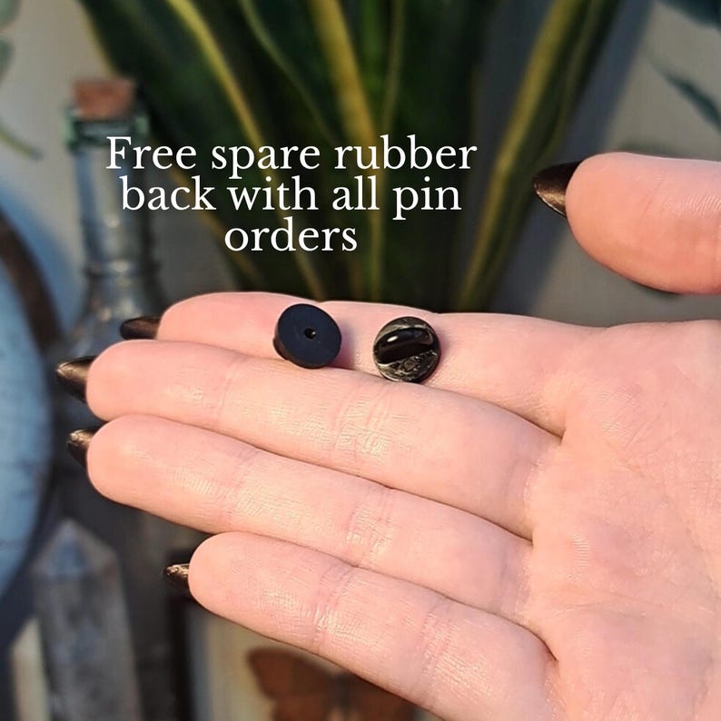May include: Close-up of two pin backs, one black rubber and one metal. The text "Free spare rubber back with all pin orders" is visible in white. The items are displayed on a hand.