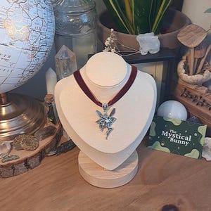 K&ouml;nnte beinhalten: Eine silberne Feen-Anh&auml;nger-Kette mit einem burgunderfarbenen Samt-Choker. Die Kette wird auf einer wei&szlig;en Mannequin-B&uuml;ste mit einem Holzsockel pr&auml;sentiert. Der Text "The Mystical Bunny" ist auf einer gr&uuml;n-wei&szlig;en Karte sichtbar.