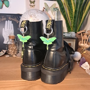 May include: Black leather boots with a green and yellow butterfly charm attached to each boot. The boots have a thick black sole and a yellow stitch around the top of the boot.
