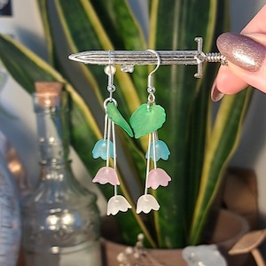 May include: A pair of dangle earrings featuring a green leaf charm and three flower charms in shades of pink, blue, and white.