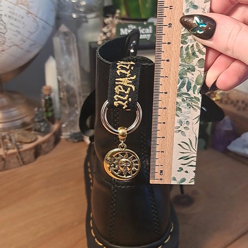 May include: Black leather boots with a gold charm featuring a sun and moon design. The boots have a black strap with gold lettering that reads "witchcraft".