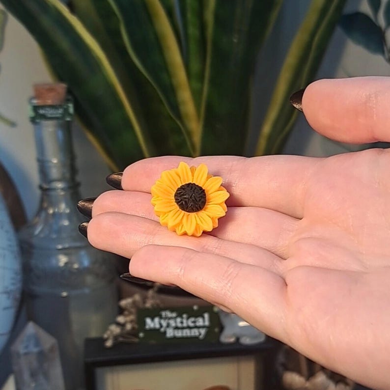 May include: A miniature sunflower with bright yellow petals and a dark brown center. The small, detailed flower is held in an open hand, showcasing its delicate design. The background includes a green plant and a glass bottle.