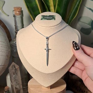 May include: A silver sword pendant necklace on a silver chain. The sword has a detailed hilt and a pointed tip. The necklace is displayed on a white mannequin bust.