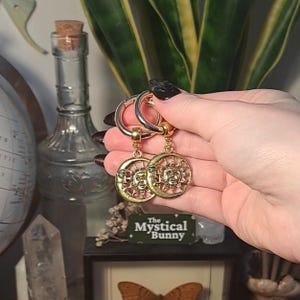 May include: Gold hoop earrings with a sun and moon charm. The sun and moon charms are detailed with faces and rays. The earrings are hanging from the hoops.