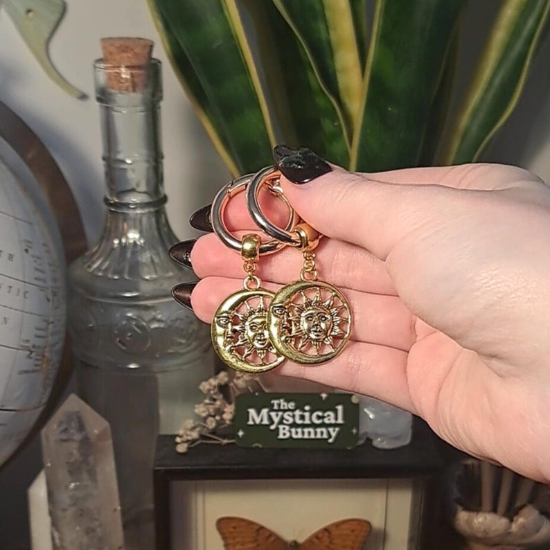 May include: Gold hoop earrings with a sun and moon charm. The sun and moon charms are detailed with faces and rays. The earrings are hanging from the hoops.