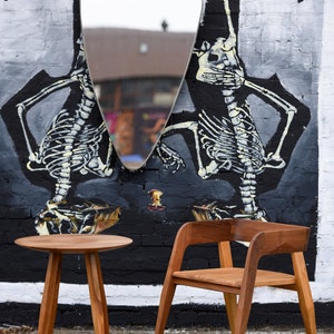 May include: A wooden table and chair set against a brick wall with a large, abstract mural of skeletons. The table has a round top and three legs, while the chair has a curved back and four legs.