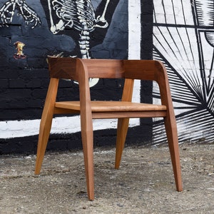 May include: A wooden chair with a brown finish and a simple design. The chair has a wide, curved back and a seat with a slight curve. The chair is sitting in front of a brick wall with a black and white mural.