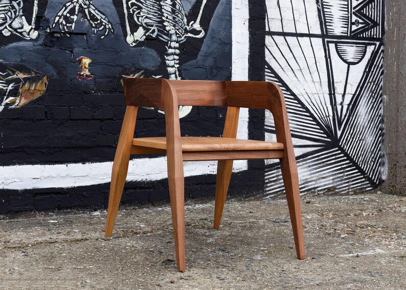 May include: A wooden chair with a brown finish and a simple design. The chair has a wide, curved back and a seat with a slight curve. The chair is sitting in front of a brick wall with a black and white mural.