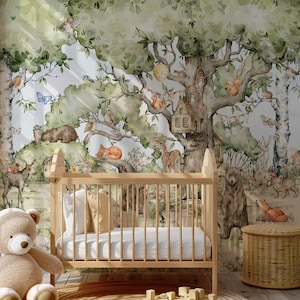 Op de afbeelding: Een babykamer met een wieg en een teddybeer. De muur is versierd met een aquarelmuurschildering van een boslandschap met dieren, waaronder herten, eekhoorns, uilen en een vos. De muurschildering toont een grote boom met een vogelhuisje en een klein huis.