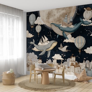 May include: A whimsical nursery wall mural featuring a dark blue night sky with white stars, clouds, and a large, full moon. Two blue whales fly through the sky with hot air balloons and ships. The mural also features a cityscape of white buildings with brown roofs.