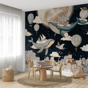 May include: A whimsical nursery wall mural featuring a dark blue night sky with white stars, clouds, and a large, full moon. Two blue whales fly through the sky with hot air balloons and ships. The mural also features a cityscape of white buildings with brown roofs.