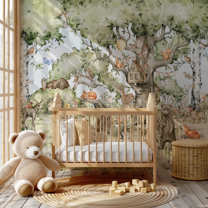 Op de afbeelding: Een babykamer met een wieg en een teddybeer. De muur is versierd met een aquarelmuurschildering van een boslandschap met dieren, waaronder herten, eekhoorns, uilen en een vos. De muurschildering toont een grote boom met een vogelhuisje en een klein huis.