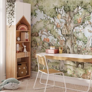 Op de afbeelding: Een wit bureau met een houten blad staat voor een muurschildering die een fantasierijk boslandschap voorstelt met een boomhut, dieren en bladeren. De muurschildering is geschilderd in aquarelstijl met tinten groen, bruin en blauw. Een witte stoel met een houten zitting staat naast het bureau.